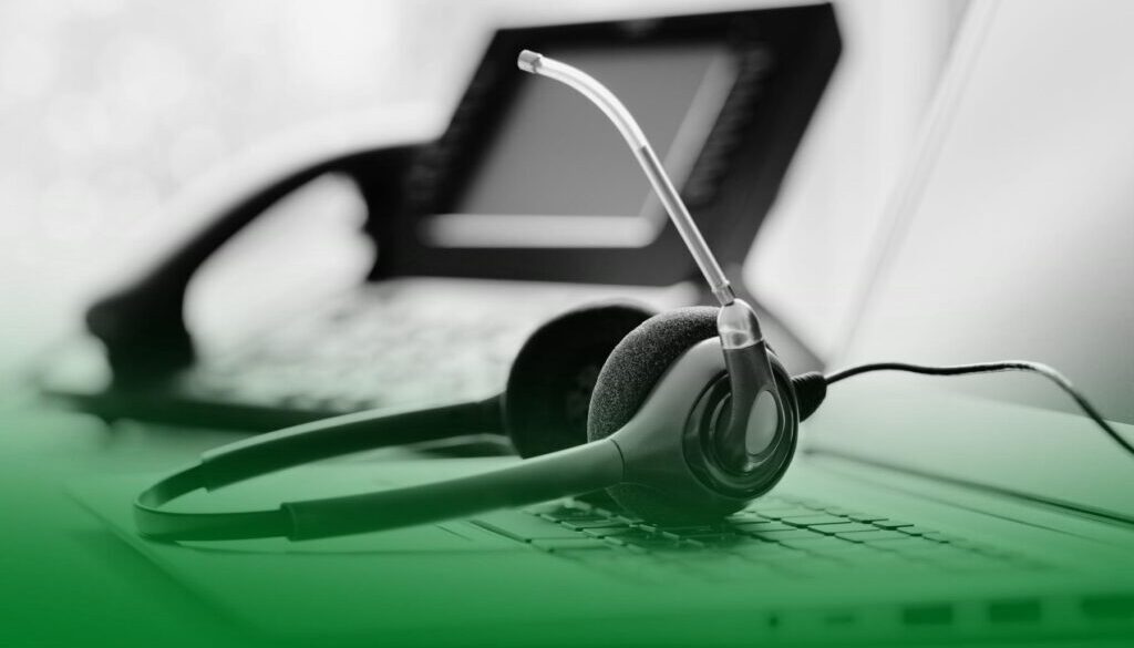 Auriculares con micrófono apoyados sobre un teclado de ordenador, con un teléfono fijo desenfocado al fondo. Auriculares con micrófono apoyados sobre un teclado de ordenador, con un teléfono fijo desenfocado al fondo.