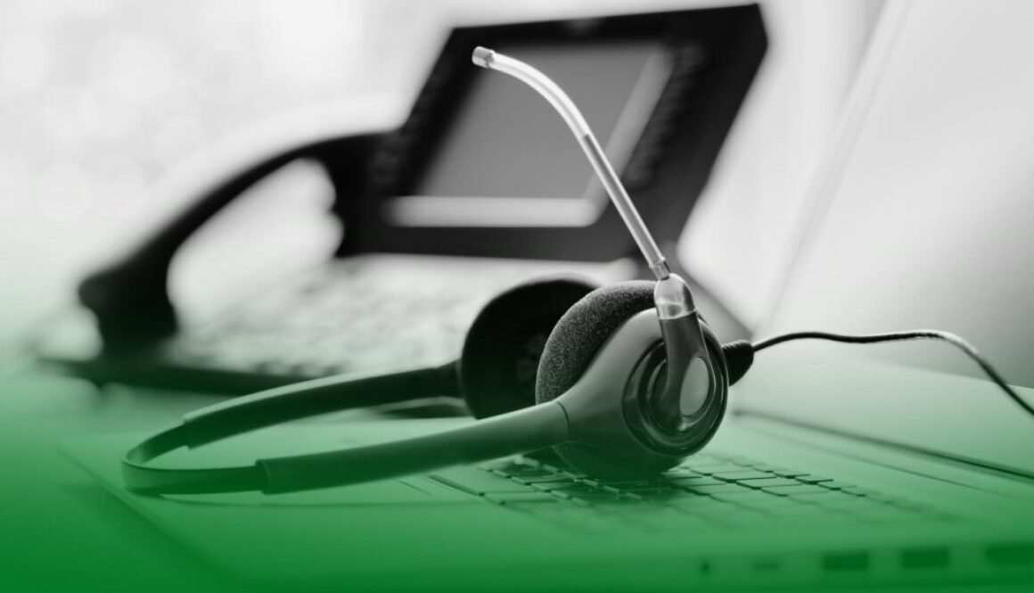 Auriculares con micrófono apoyados sobre un teclado de ordenador, con un teléfono fijo desenfocado al fondo. Auriculares con micrófono apoyados sobre un teclado de ordenador, con un teléfono fijo desenfocado al fondo.