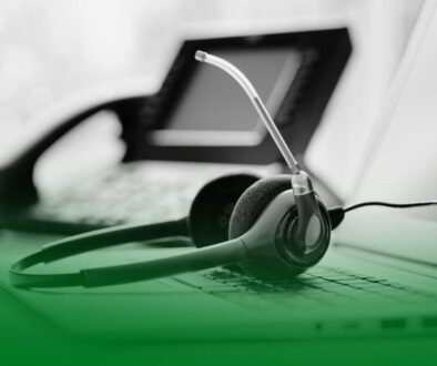 Auriculares con micrófono apoyados sobre un teclado de ordenador, con un teléfono fijo desenfocado al fondo. Auriculares con micrófono apoyados sobre un teclado de ordenador, con un teléfono fijo desenfocado al fondo.