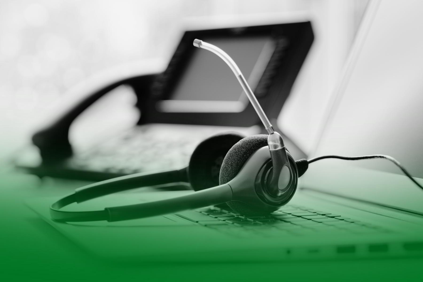 Auriculares con micrófono apoyados sobre un teclado de ordenador, con un teléfono fijo desenfocado al fondo. Auriculares con micrófono apoyados sobre un teclado de ordenador, con un teléfono fijo desenfocado al fondo.