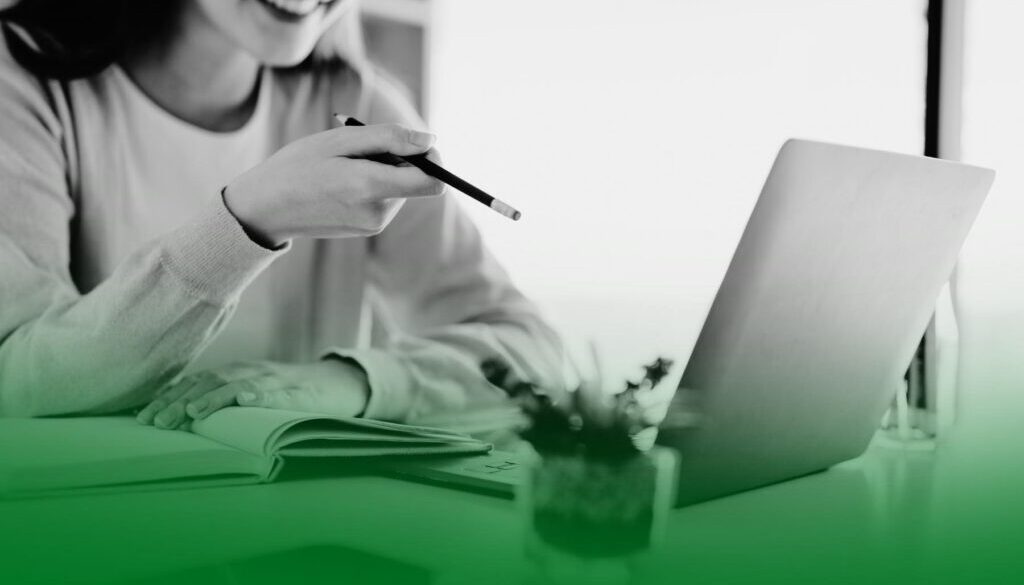 Mujer sonriendo frente a un portátil mientras señala la pantalla con un bolígrafo y toma notas en una libreta. Mujer sonriendo frente a un portátil mientras señala la pantalla con un bolígrafo y toma notas en una libreta.