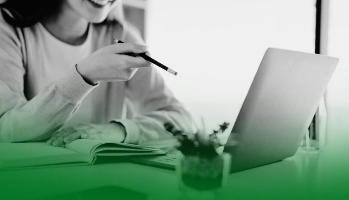 Mujer sonriendo frente a un portátil mientras señala la pantalla con un bolígrafo y toma notas en una libreta. Mujer sonriendo frente a un portátil mientras señala la pantalla con un bolígrafo y toma notas en una libreta.