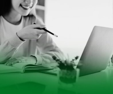 Mujer sonriendo frente a un portátil mientras señala la pantalla con un bolígrafo y toma notas en una libreta. Mujer sonriendo frente a un portátil mientras señala la pantalla con un bolígrafo y toma notas en una libreta.