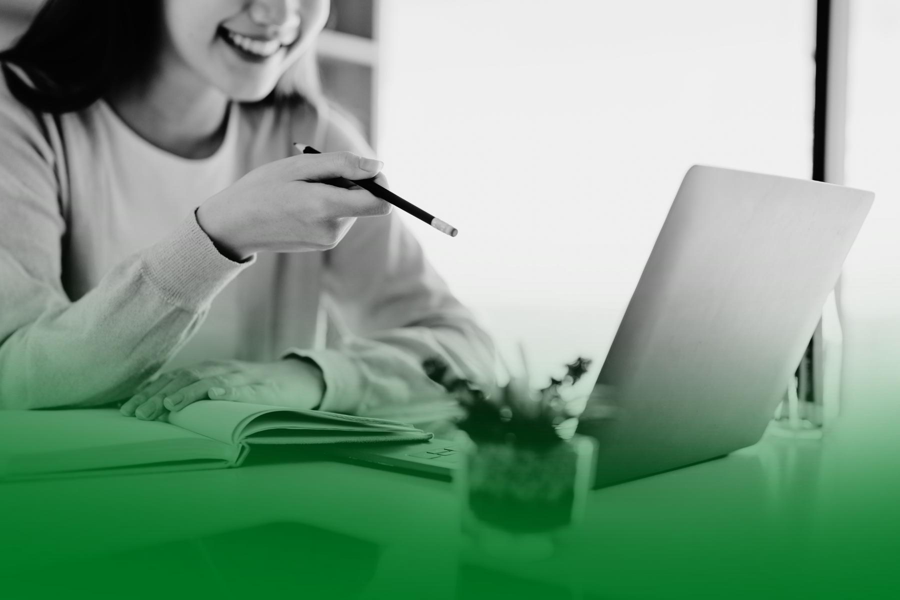 Mujer sonriendo frente a un portátil mientras señala la pantalla con un bolígrafo y toma notas en una libreta. Mujer sonriendo frente a un portátil mientras señala la pantalla con un bolígrafo y toma notas en una libreta.