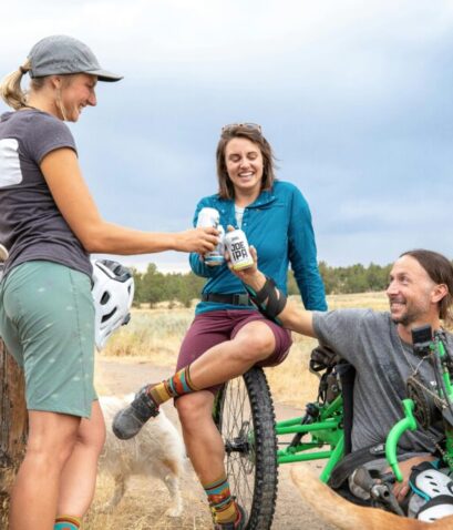 Tres personas ciclistas, una de ellas en bicicleta adaptada, brindando y sonriendo en un descanso